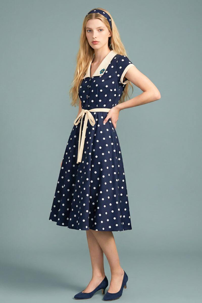 A side view of a model wearing a navy polka dot midi dress with an ivory collar and waist tie, showing the fluid skirt movement and classic feminine silhouette.