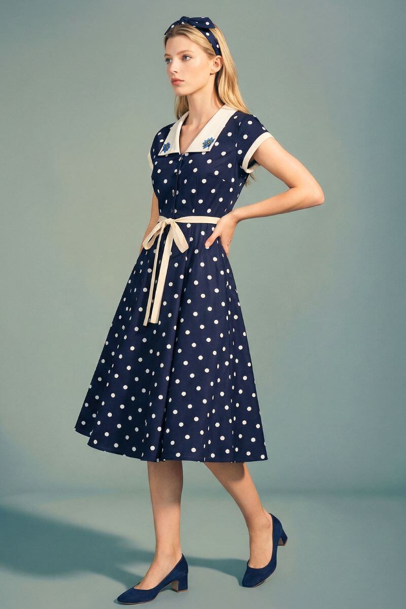 A model wears a navy polka dot midi dress with a contrast ivory collar, button-front bodice, and tied waist belt, styled with matching navy heels against a soft neutral studio background.