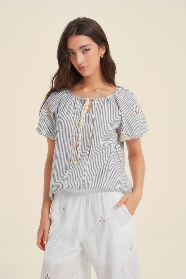 Front view of a woman in a navy and white striped blouse featuring lace shoulder inserts, puff sleeves, and tasseled neckline ties, styled with white embroidered pants.