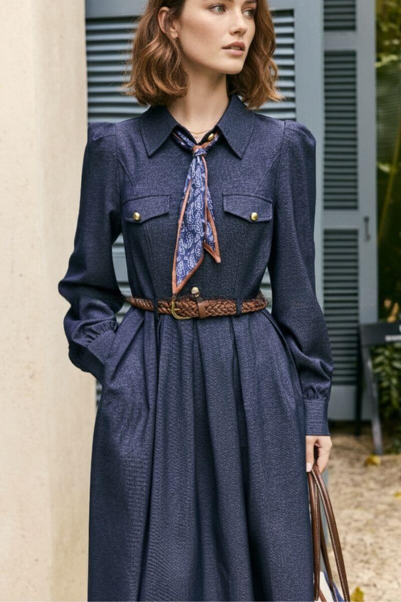 Model standing outdoors wearing a navy twill midi dress with long sleeves, gold button chest pockets, a brown braided belt, and a paisley neck scarf, styled with a structured handbag.