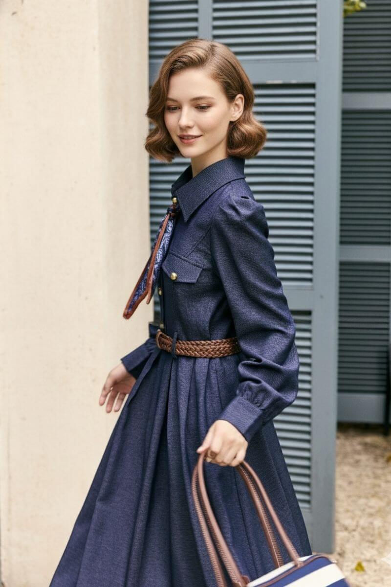 Model walking outdoors wearing a navy twill belted midi dress featuring gold buttons, puff sleeves, and a brown woven belt, styled with a blue paisley neck scarf and striped handbag.