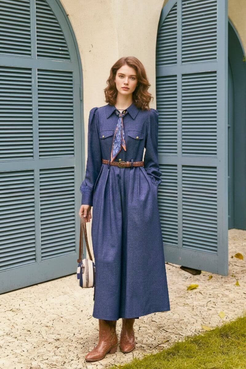 Model standing in front of blue shutters wearing a navy twill long sleeve belted midi dress with gold buttons, paisley neck scarf, and brown boots, holding a striped handbag.