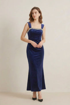 A woman wearing a navy velvet maxi dress with pearl embellishment along the square neckline, standing in a minimal studio environment.