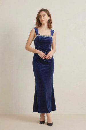 A woman wearing a navy velvet maxi dress with pearl embellishment along the square neckline, standing in a minimal studio environment.