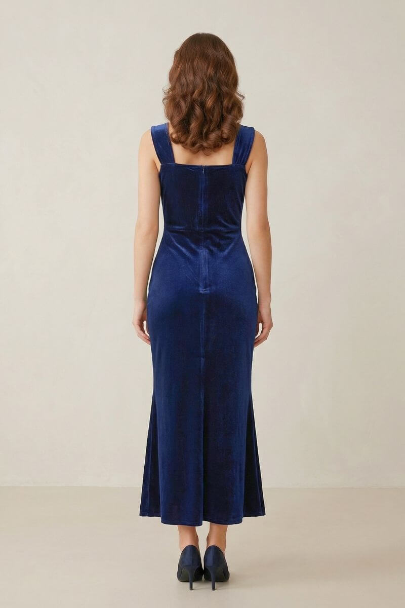 A woman wearing a navy velvet square-neck maxi dress viewed from the back, showing its smooth fitted silhouette and wide straps in a soft studio setting.