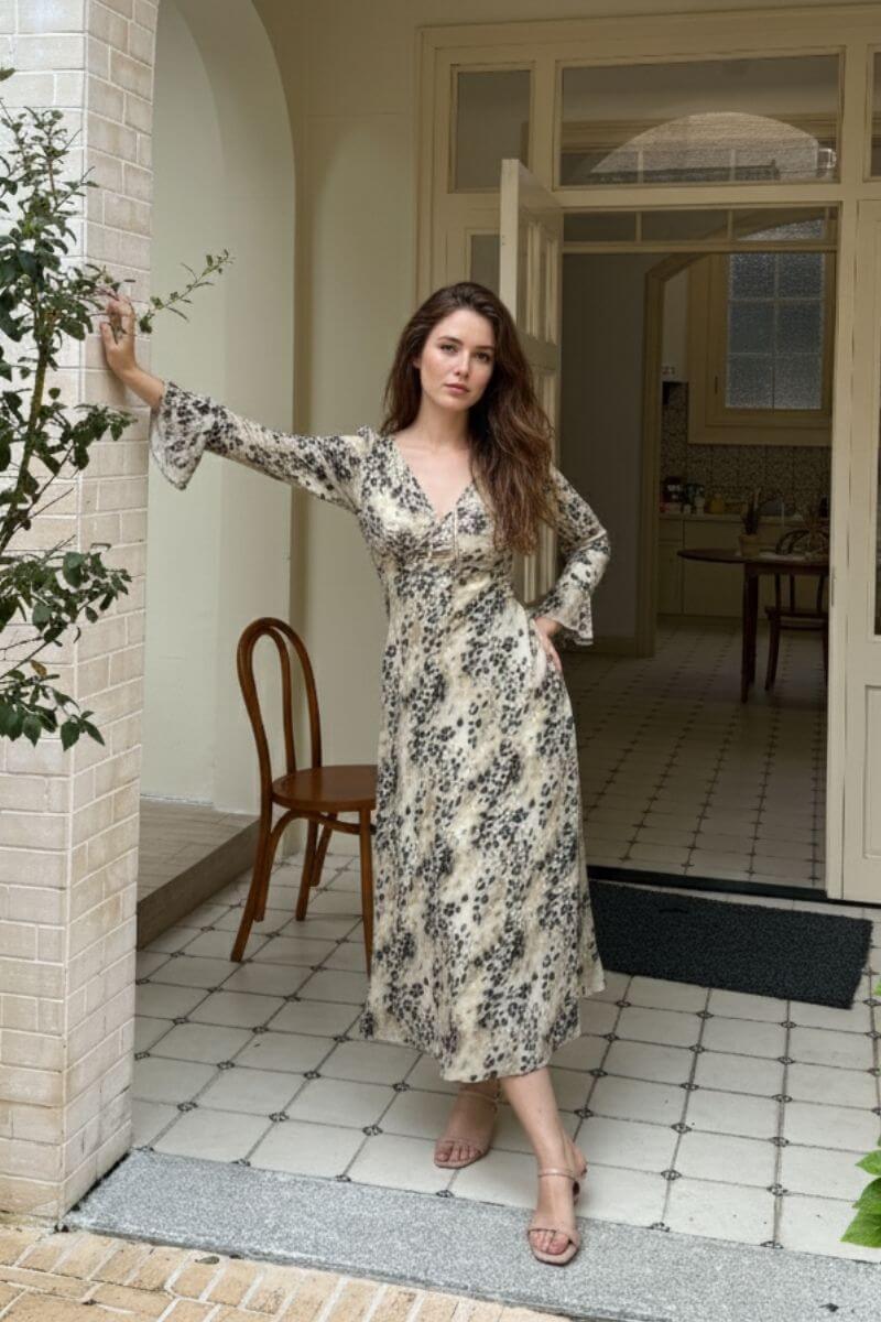 Model posing near a tiled entrance wearing a neutral beige and black leopard print midi dress with long fluted sleeves and soft draping, styled with nude heels for an effortlessly elegant daytime look.