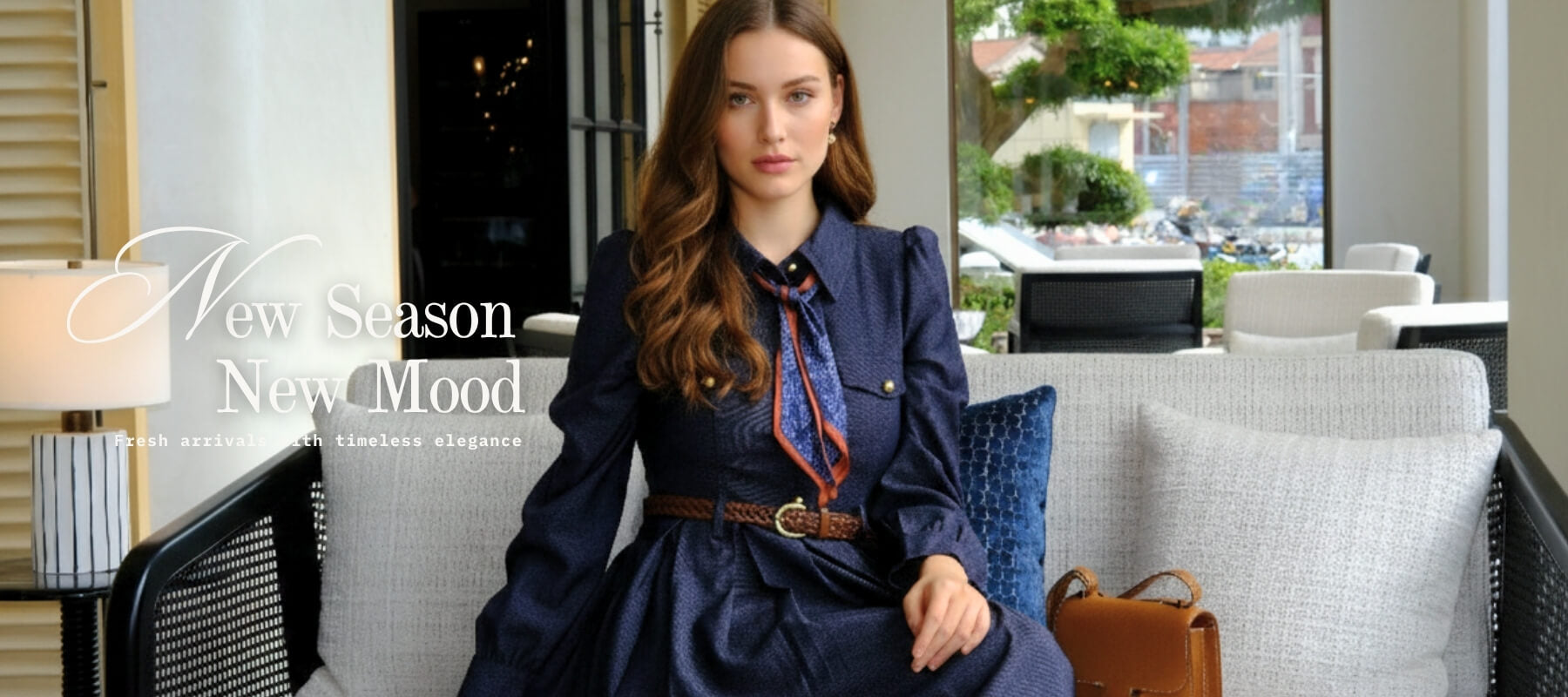 Woman seated on a modern sofa wearing a navy long-sleeve belted midi dress with a patterned scarf tie, styled with soft waves and a brown leather bag beside her — part of Sioéne’s “New Season, New Mood” campaign.
