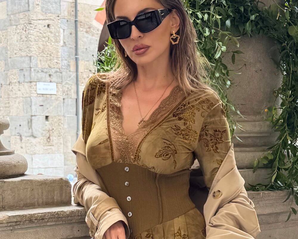 A woman wearing an olive green velvet floral corset maxi dress with lace trim and long sleeves, styled with a beige trench coat and black sunglasses, standing in a European stone courtyard. A vintage-inspired elegant fall outfit by Sioéne.