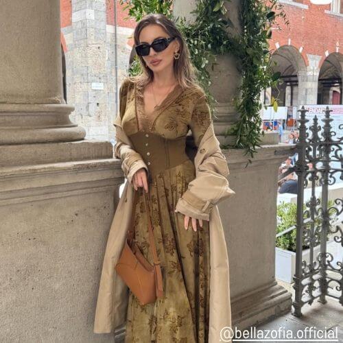 Woman wearing an olive green velvet corset maxi dress with a beige trench coat and brown handbag from Sioéne, posing in a European-style street scene — elegant travel outfit inspiration.