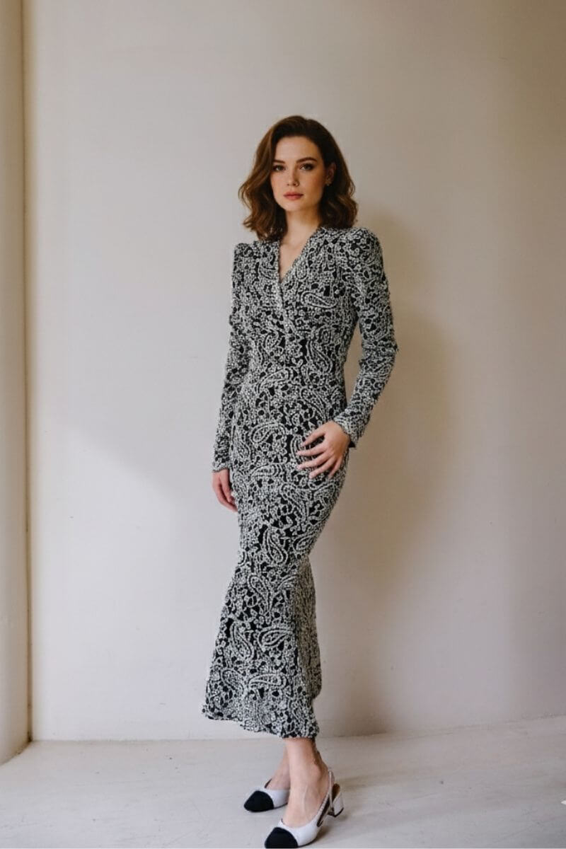 Model posing against a neutral wall in a fitted black and white paisley print midi dress with puff shoulders and a flared hem, paired with cream and black slingback heels for a refined vintage look.