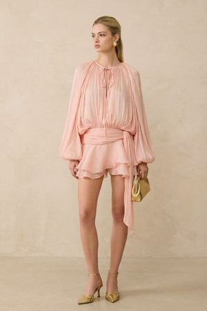 Model wears a soft peach chiffon blouson top with gathered neckline and layered ruffle shorts, styled with gold heels and a mini clutch against a warm neutral backdrop.