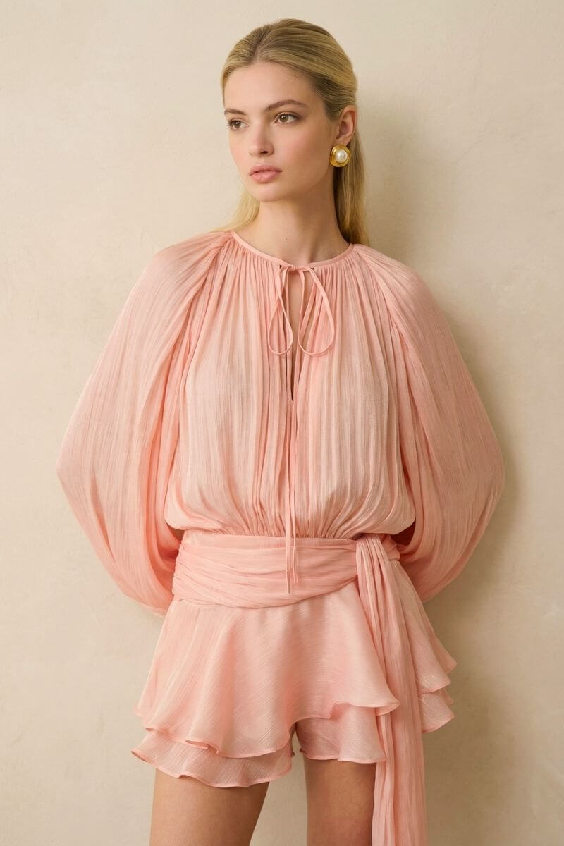 Close-up of a peach chiffon blouson top featuring fine pleats, a delicate tie neckline, and airy long sleeves with a softly cinched waist.