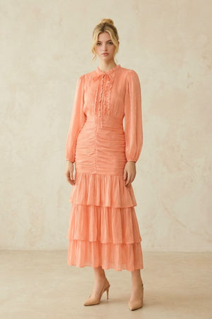 Model stands slightly angled wearing a peach chiffon midi dress featuring gathered ruching, sheer sleeves, and layered ruffle hem for a romantic silhouette.