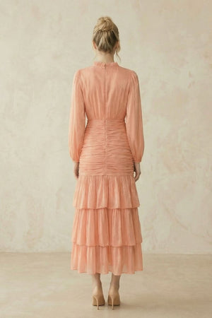 Back view of a peach midi dress with sheer long sleeves, ruched waist detailing, and a softly tiered skirt, shown on a model with hair in a low bun.