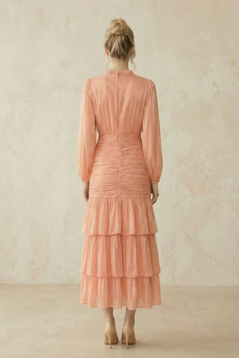 Back view of a peach midi dress with sheer long sleeves, ruched waist detailing, and a softly tiered skirt, shown on a model with hair in a low bun.