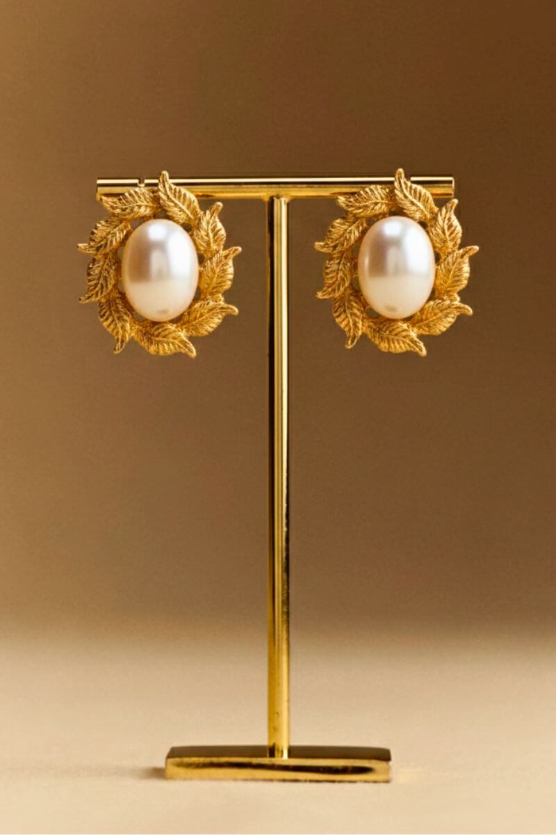 Gold leaf stud earrings with oval pearls displayed on a minimalist jewelry stand against a warm brown backdrop, highlighting their sculptural vintage-inspired design.
