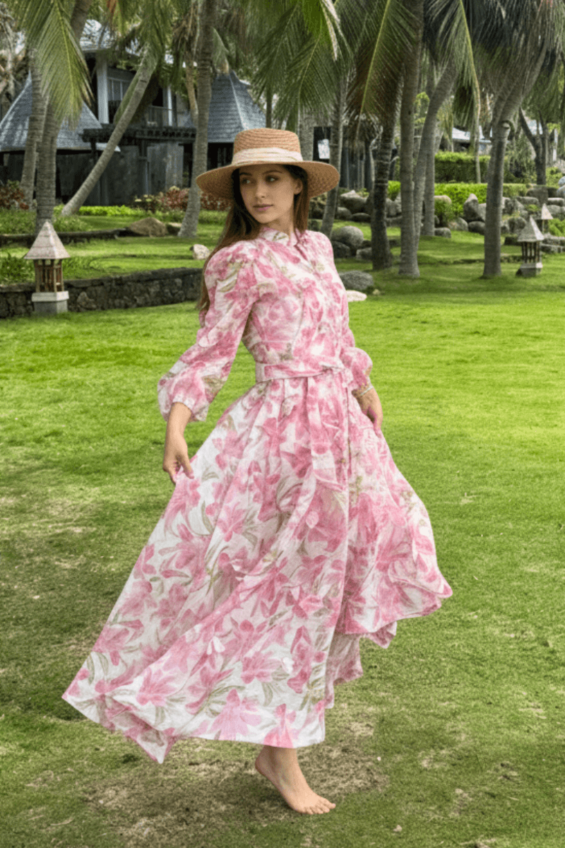 Woman in a pink floral maxi dress with long sleeves and a waist tie, barefoot and twirling gracefully in a tropical garden.