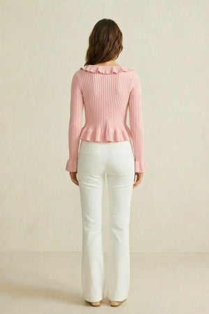 Back view of a woman in a pink ribbed knit top with ruffled neckline and hem, styled with white flared pants.