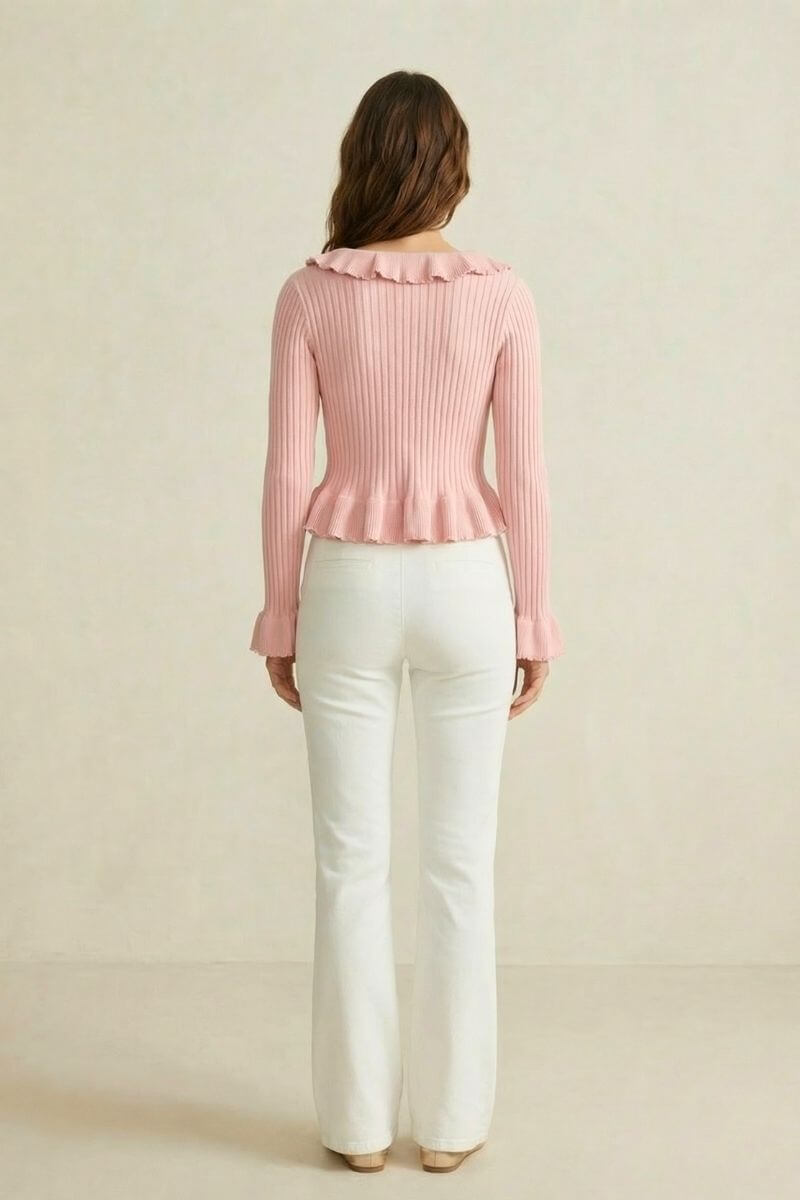 Back view of a woman in a pink ribbed knit top with ruffled neckline and hem, styled with white flared pants.
