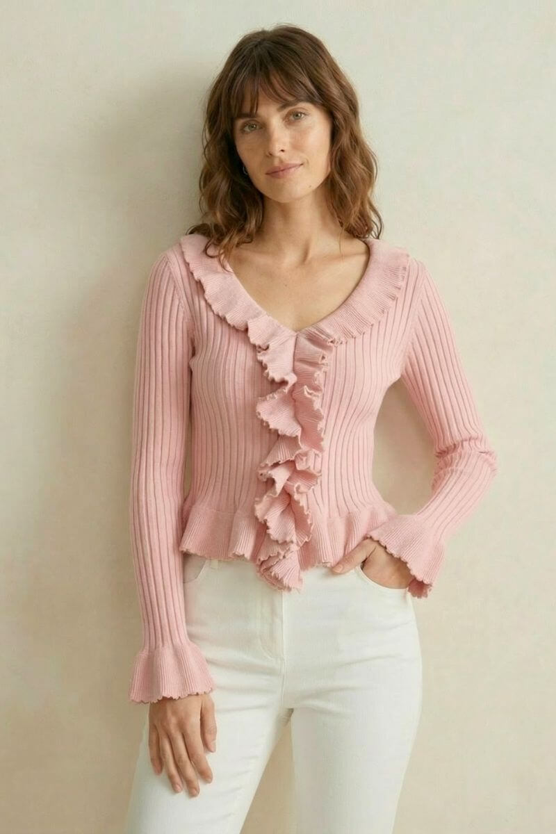 Woman wearing a pink ribbed knit top featuring cascading front ruffles and flared sleeves, paired with white pants against a soft neutral background.