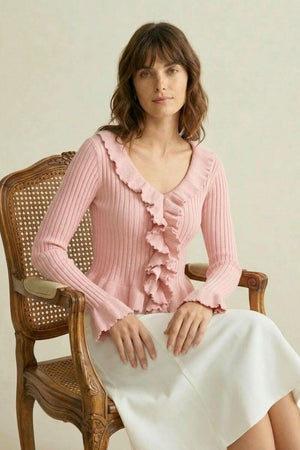 Woman sitting on a vintage wooden chair wearing a pink ribbed knit top with ruffle trim and a white skirt, soft neutral background.