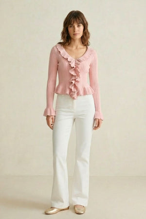 Woman standing in a pink ribbed knit ruffle top styled with white flared pants and gold flats against a neutral studio backdrop.