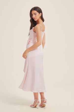 Woman posing sideways in a pink satin slip midi dress featuring a side slit and floral detailing at the waist, styled with white strappy heels.