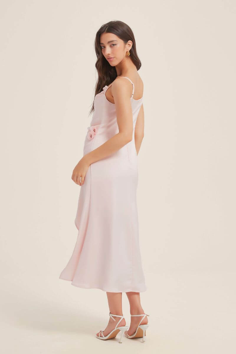 Woman posing sideways in a pink satin slip midi dress featuring a side slit and floral detailing at the waist, styled with white strappy heels.