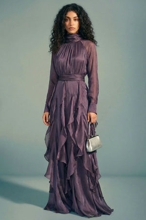 Model facing forward in a plum satin maxi dress featuring a softly gathered high neckline, cinched waist, sheer long sleeves, and fluid ruffled skirt with a subtle sheen.