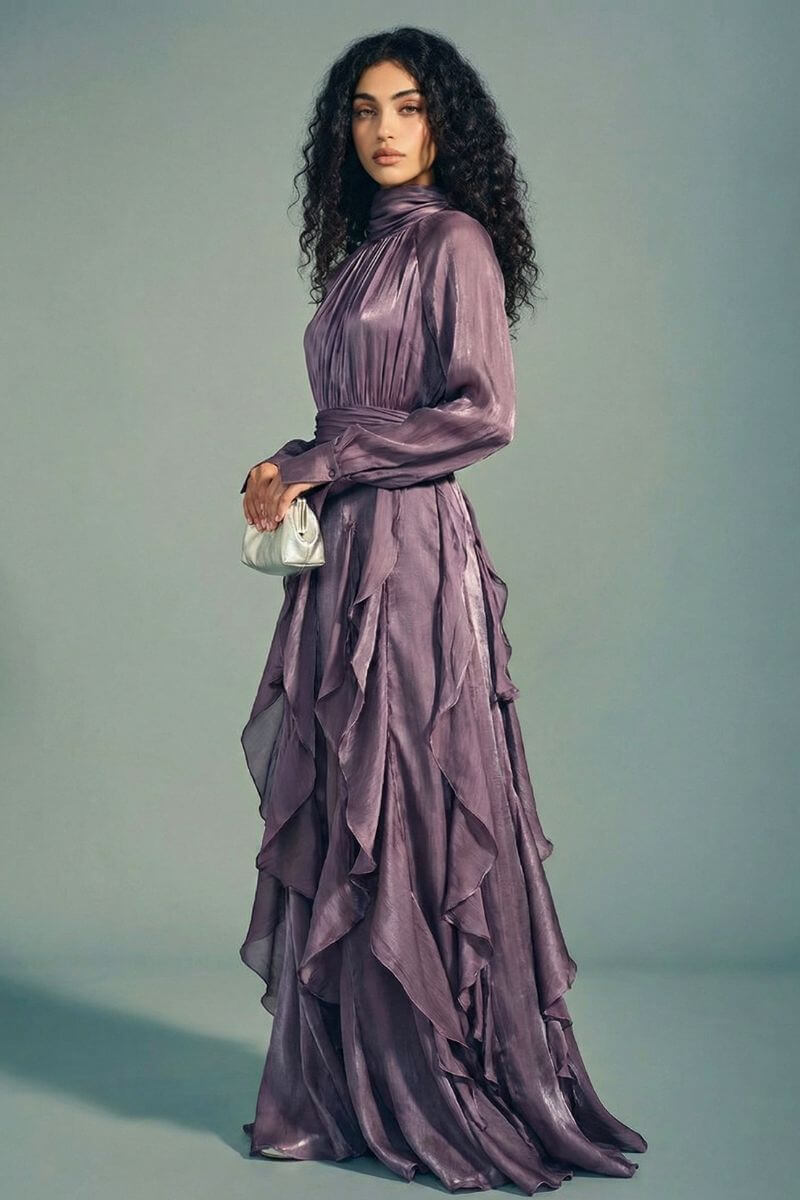 Model standing in a side pose wearing a plum satin maxi dress with a high gathered neckline, sheer long sleeves, and cascading ruffle layers, styled with a small metallic clutch.