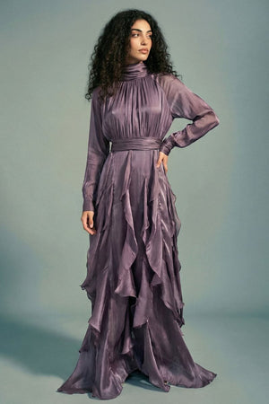 Model turned slightly to the side wearing a plum satin maxi dress with a defined waist, elegant drape, sheer sleeves, and layered ruffle detailing flowing down the skirt.