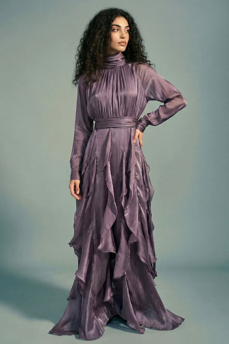 Model turned slightly to the side wearing a plum satin maxi dress with a defined waist, elegant drape, sheer sleeves, and layered ruffle detailing flowing down the skirt.