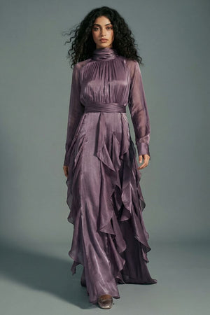Model walking forward in a plum satin maxi dress with a softly structured high neck, sheer long sleeves, cinched waist, and airy ruffle layers that move fluidly with each step.