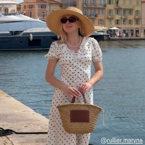 Woman wearing a white polka-dot midi dress from Sioéne with a straw hat and woven tote, standing by a yacht harbor under sunlight — vintage summer outfit inspiration.
