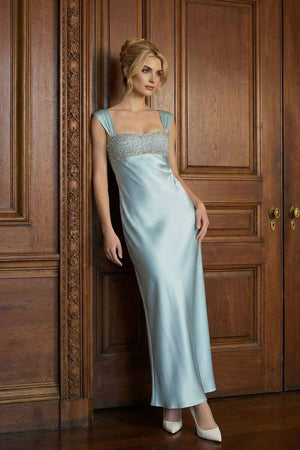 A model leans against a carved wooden door wearing a powder blue satin maxi dress with wide straps and a beaded straight neckline, styled with ivory heels in a classic interior setting.