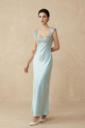 A model poses at a slight angle wearing a powder blue satin maxi dress with a softly fitted waist, beaded neckline detail, and smooth drape through the skirt.