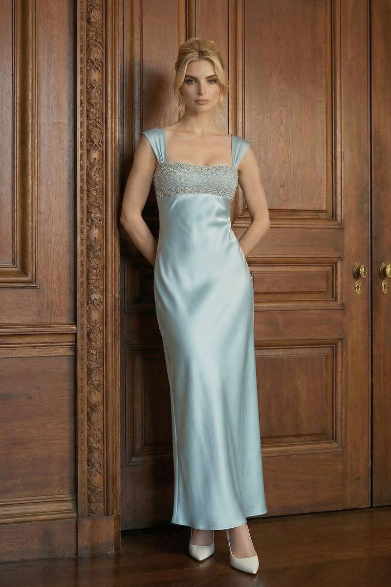A model stands against a wooden paneled wall wearing a powder blue satin maxi dress with a beaded neckline, wide straps, and a fluid, elegant skirt.