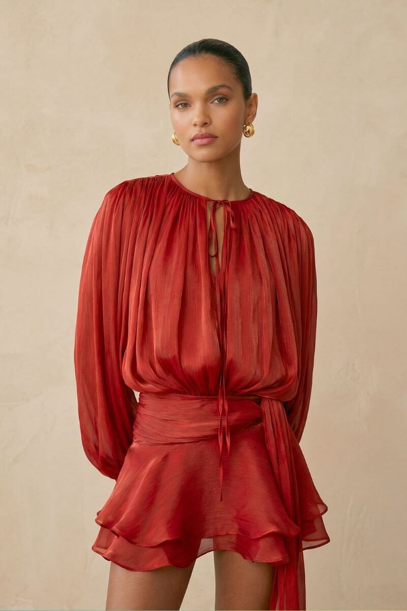 A close view of the red chiffon mini dress highlighting the pleated neckline with a delicate tie detail, sheer long sleeves, and fluid texture that drapes softly over the body.
