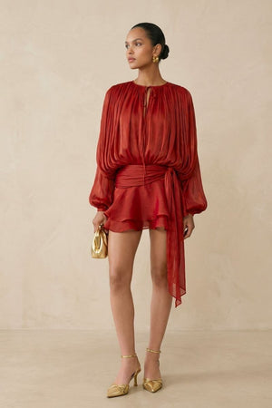 A model wears a deep red chiffon long-sleeve mini dress with a softly gathered neckline, cinched waist sash, and layered ruffle hem, styled with gold heels against a warm neutral studio backdrop.