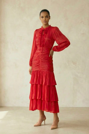 A full-body view of a woman in a red chiffon long-sleeve midi dress featuring a ruched waist and layered tiered skirt, styled with nude heels in a minimal studio.