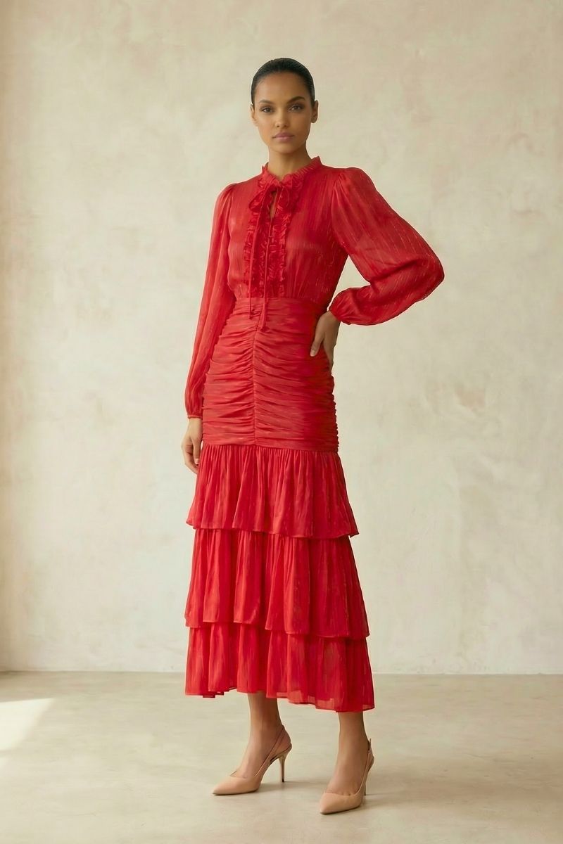 A full-body view of a woman in a red chiffon long-sleeve midi dress featuring a ruched waist and layered tiered skirt, styled with nude heels in a minimal studio.