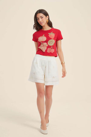 Woman wearing a red fruit print graphic crop tee with strawberry, kiwi, and cherry illustrations, styled with white embroidered shorts and white flats for a playful summer look.