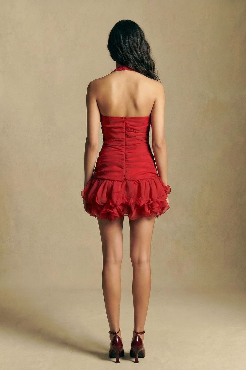 The back view highlights the red halter mini dress with an open back, fitted bodice, and softly layered rosette hem, captured in a neutral-toned studio.
