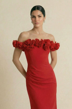 A model wearing an off-shoulder red gown adorned with sculpted rosette trim across the neckline, posing against a neutral studio background in an elegant, refined look.