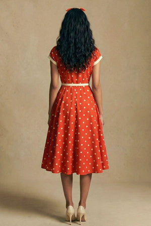 Model seen from the back wearing a red polka dot midi dress with a softly flared skirt, cream trim details, and a fitted waist, styled with neutral heels against a warm studio backdrop.