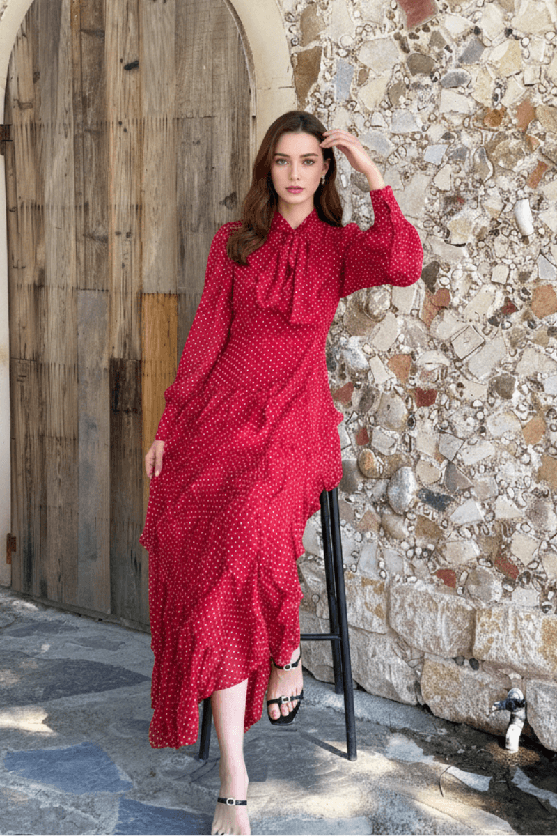 A woman sits on a tall stool against a stone wall, wearing a red long-sleeve polka dot maxi dress with a bow neckline and ruffled hem.