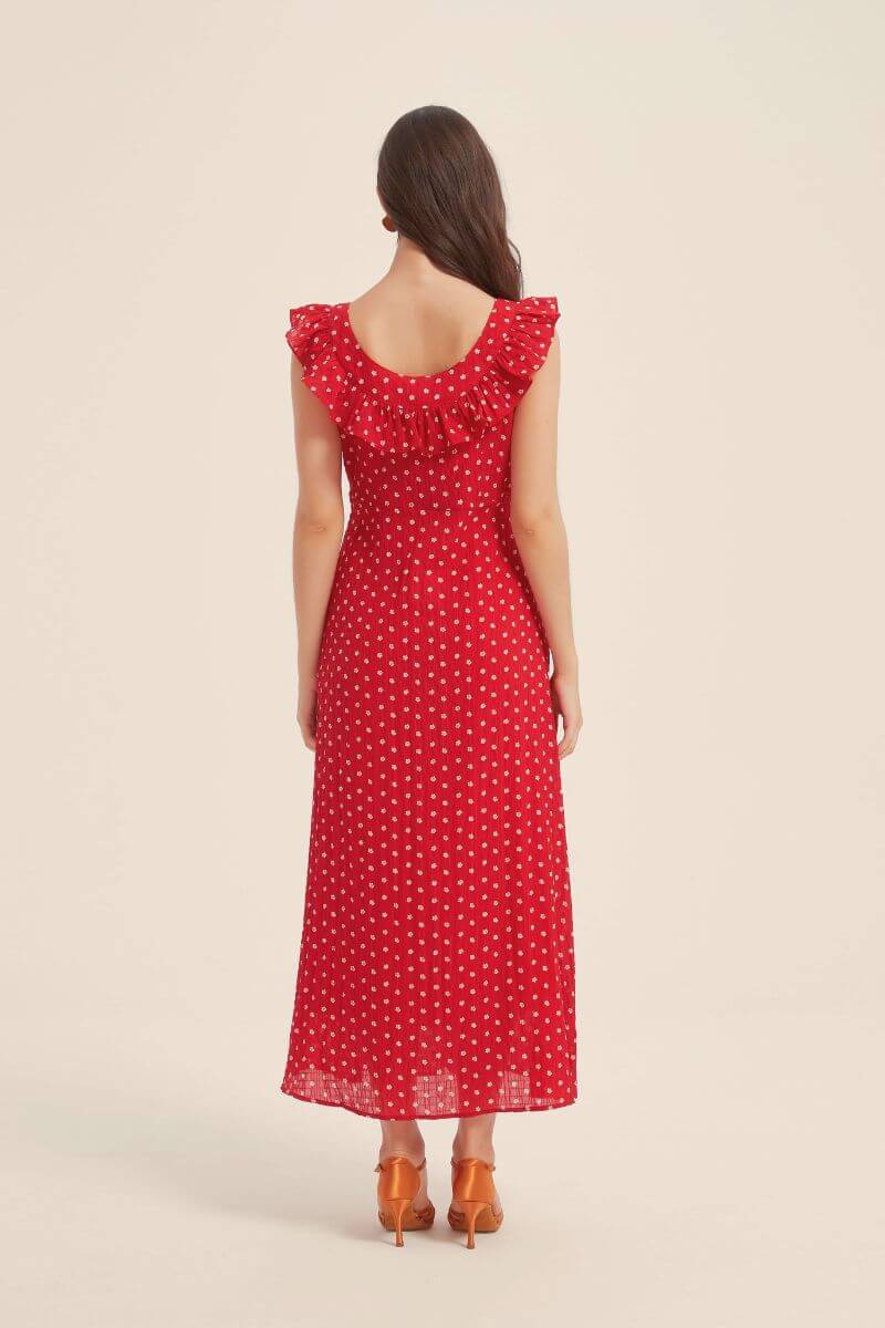 Back view of a woman wearing a red sleeveless maxi dress with white polka dots and ruffled neckline, styled with orange heels against a neutral background.