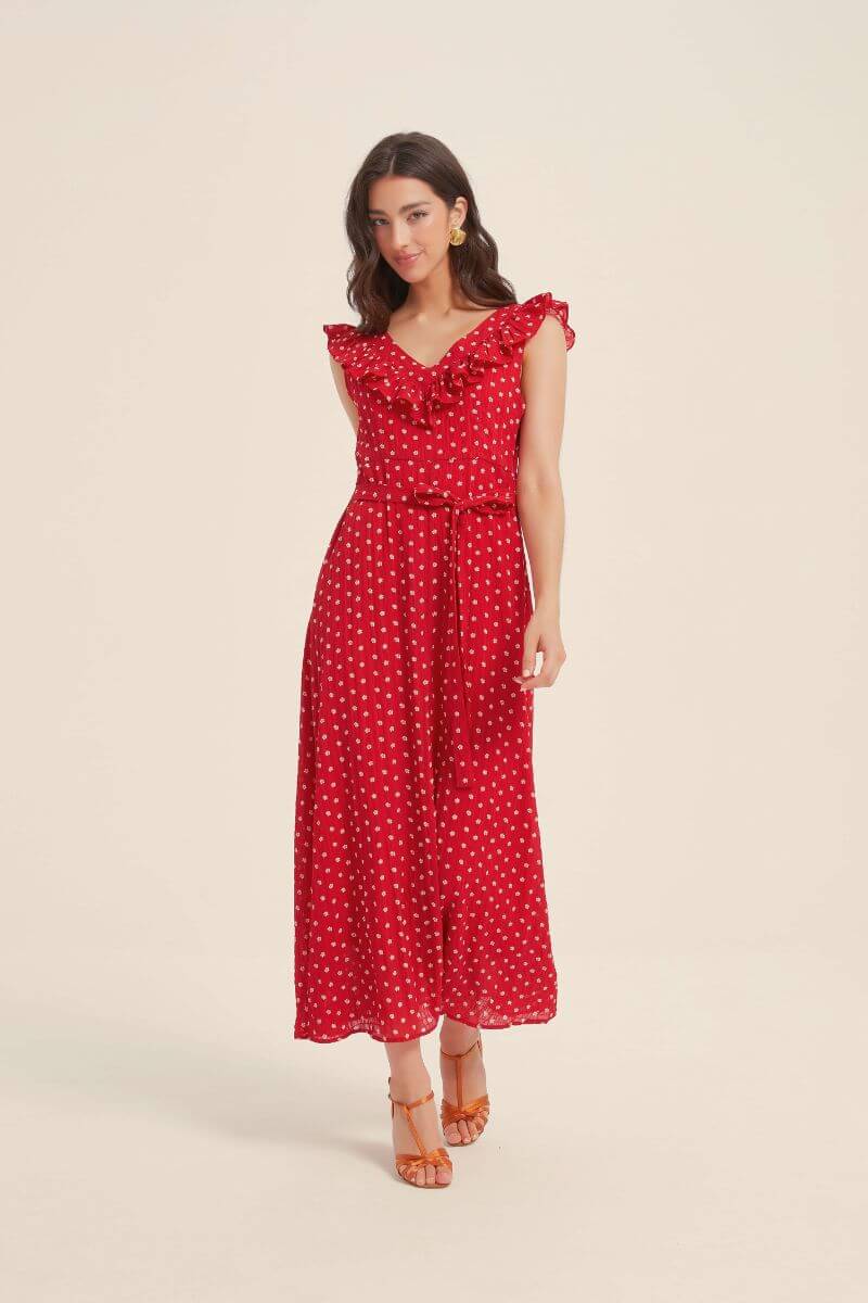 Full-length image of a woman wearing a red ruffle V-neck polka dot maxi dress with a waist tie, paired with orange heels and gold earrings on a beige backdrop.