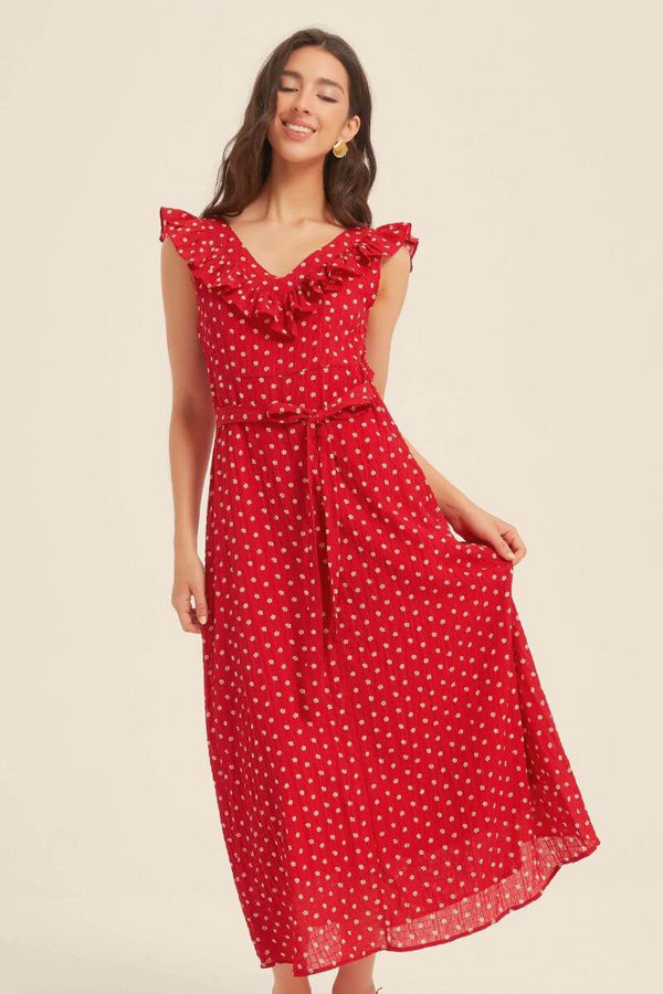 Woman smiling while wearing a red polka dot midi dress with ruffled neckline and tied waist, styled with minimal accessories against a light background.