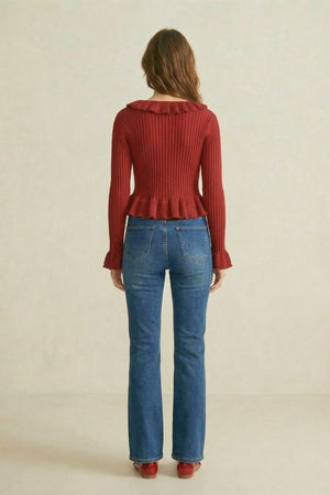 Back view of a woman wearing a red ribbed knit top with ruffle trim paired with blue jeans, neutral background.